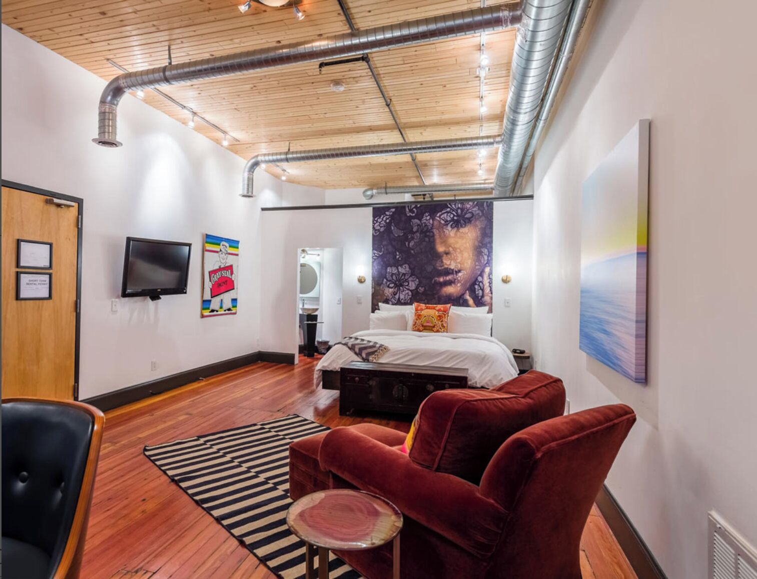 A modern bedroom with a wooden ceiling, a bed with a headboard, a red armchair, a small table, and a TV mounted on the wall.
