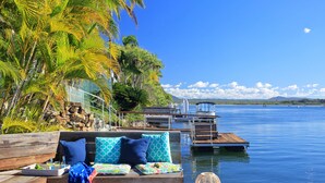 Une terrasse en bois avec un banc et des coussins, donnant sur une étendue d’eau calme, avec un quai et un petit bateau.