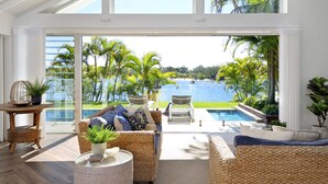 Une pièce spacieuse dotée d’une grande porte vitrée donnant sur un espace piscine, de meubles en osier et offrant une vue sur un paysage tropical.