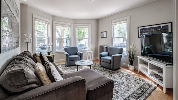 Un salon moderne avec un canapé gris, deux fauteuils, une table basse et un téléviseur posé sur un meuble blanc. De grandes fenêtres laissent entrer la lumière naturelle et offrent une vue sur la rue.