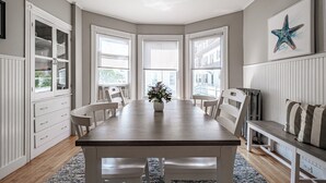 Un espace repas comprenant une table en bois, des chaises blanches, un banc et une vue dégagée grâce à de grandes fenêtres.