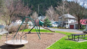 Ein Spielplatz mit Schaukeln und Rutsche, umgeben von einer Wiese und einem bewaldeten Hügel im Hintergrund.