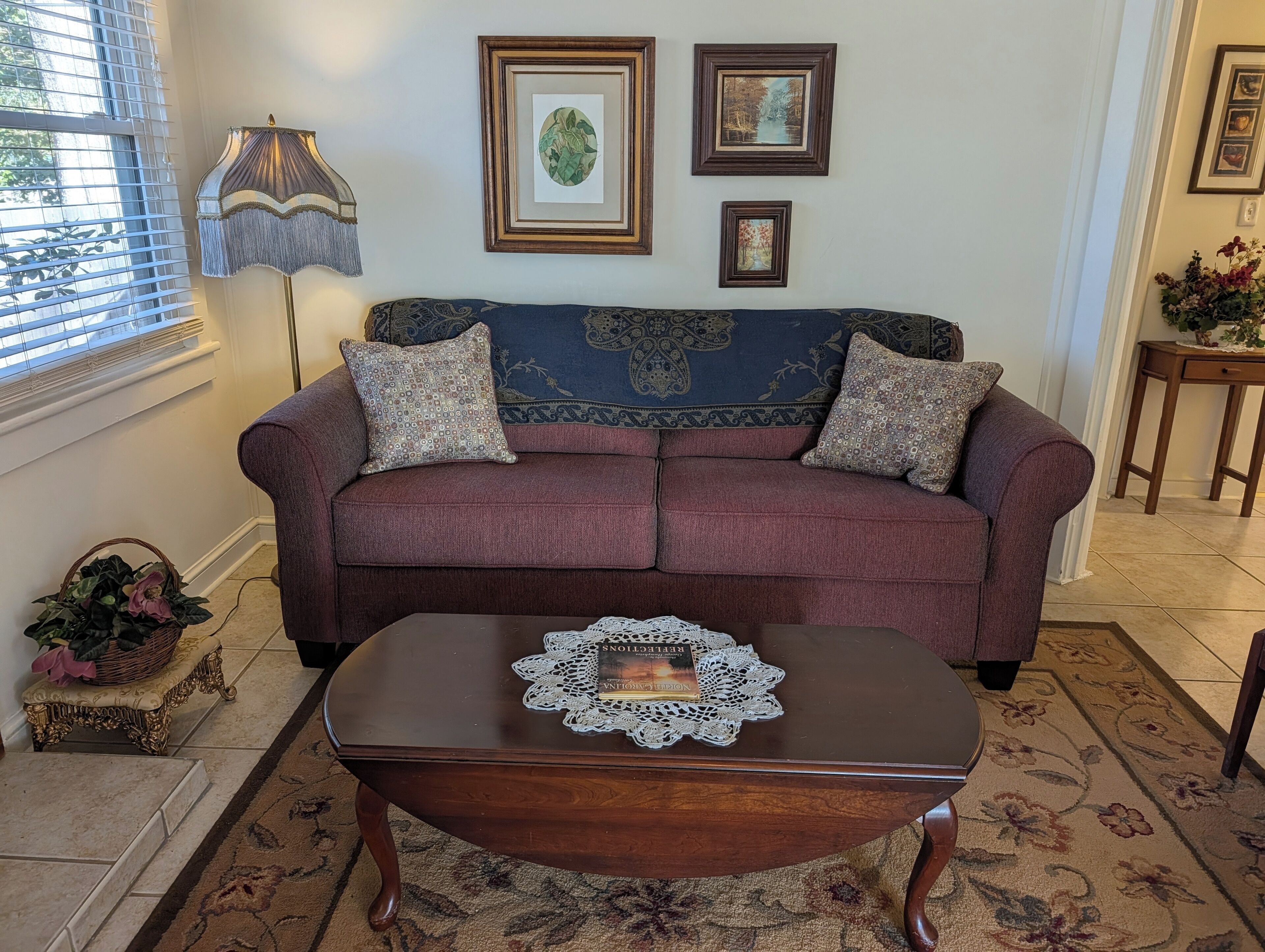 A living room with a sofa, a coffee table, and a lamp. There are framed pictures on the wall and a window with blinds.