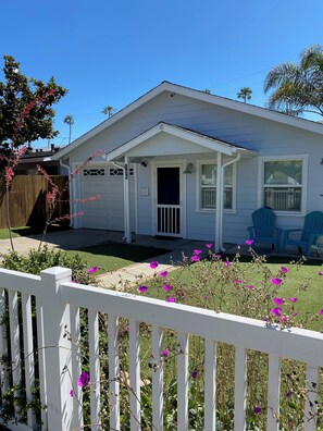 A single-story house with a front porch, a garage, and a small lawn area with purple flowers.