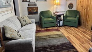 A cozy living room with a grey sofa, two green armchairs, a wooden floor, and various wall decorations including framed pictures and a lamp. - Jackson NH Peaceful, Country Cottage / Access to Fabulous Amenities (Bartlett)