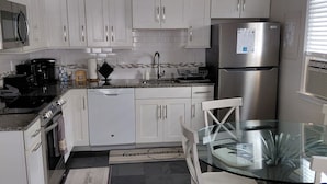 Cozinha moderna com armários brancos, geladeira de aço inoxidável, mesa de jantar de vidro e piso ladrilhado preto.