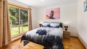 A bedroom with a large window, a bed with a forest print bedspread, two bedside tables with lamps, and a wall-mounted picture of a bird. - PET FRIENDLY COTTAGES - KELPIE COTTAGE - SELF CONTAINED COTTAGES (Worrolong, Mount Gambier)