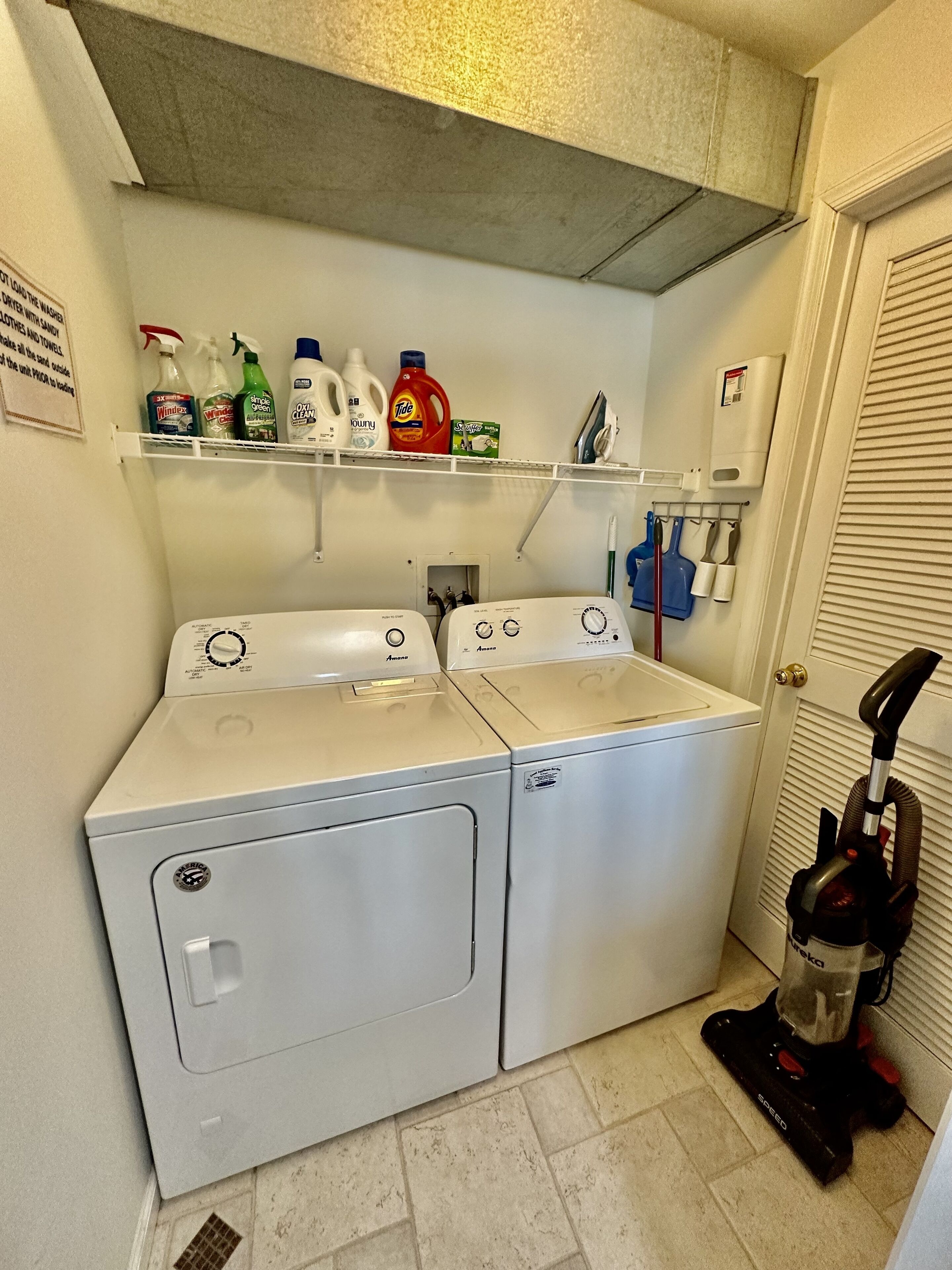 A laundry room with two front-loading washing machines, a shelf with various cleaning products, and a vacuum cleaner.