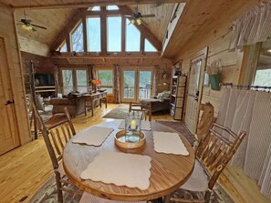 A rustic cabin interior with a wooden dining table, chairs, and a large window overlooking a deck. - River Magic, Romantic Luxury Log Cabin with Hot Tub (Hot Springs)