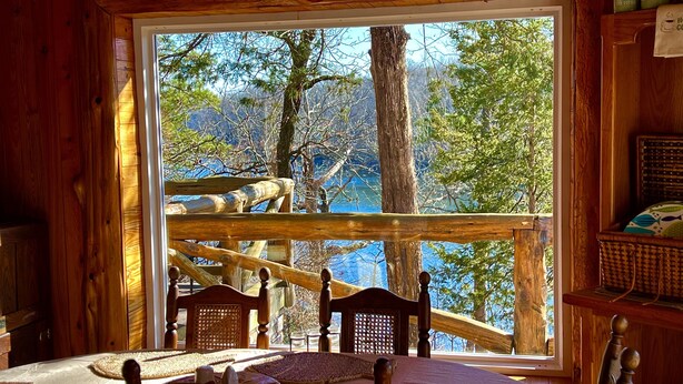 Uma cabana de madeira com uma janela grande que dá para um lago e árvores. No interior, há uma mesa com toalha branca e dois jogos americanos de palha.