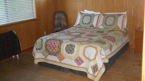 Une chambre avec un plancher en bois, un lit recouvert d’une courtepointe à motifs, une chaise et une fenêtre avec des stores.