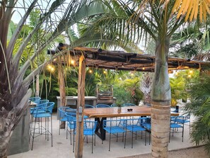 Ein Außenbereich mit einer Holztischgruppe und blauen Stühlen, umgeben von Palmen und einer Holzpergola mit Lichterketten.