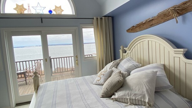 A bedroom with a bed, pillows, a wooden headboard, a view of the sea through a large window, and a balcony with a table and chairs. - Waterfront, beautiful home, breathless sunsets on Prudence Island (Prudence Island)