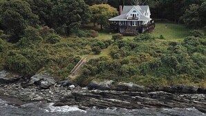 A large house surrounded by dense forest, with a rocky shoreline in the foreground. - Waterfront, beautiful home, breathless sunsets on Prudence Island (Prudence Island)