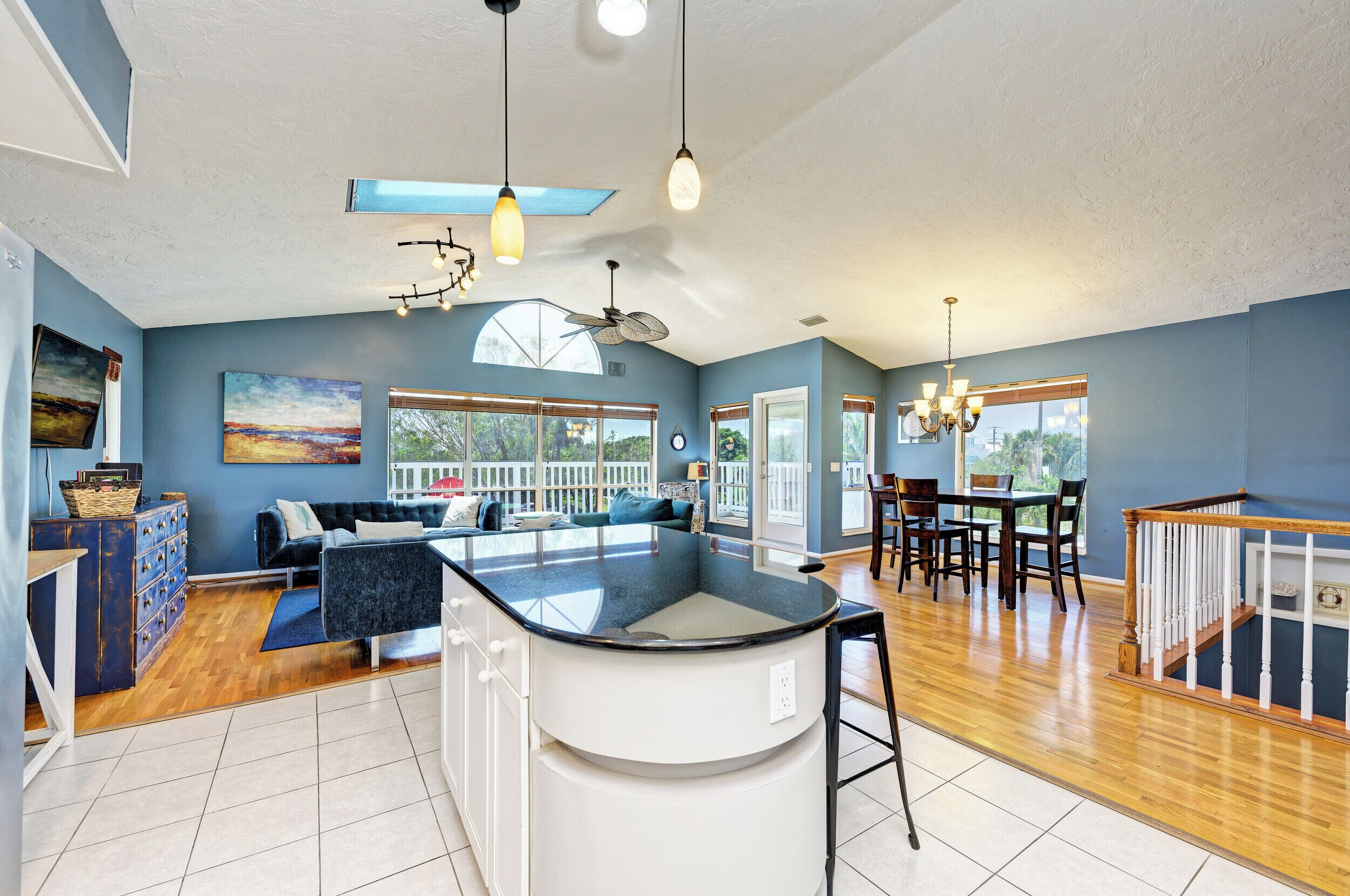 A spacious kitchen with a central island, a dining area with wooden chairs, and a living room with a sofa and a large window.