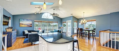 A spacious kitchen with a central island, a dining area with wooden chairs, and a living room with a sofa and a large window.