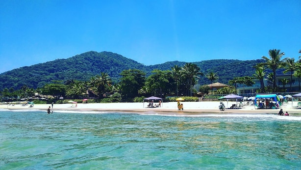 A beach with clear turquoise water, white sand, and palm trees. There are beach umbrellas and a few people in the water. - JUQUEHY MIDDLE OF THE BEACH, 80 METERS FROM THE SEA. (São Sebastião)
