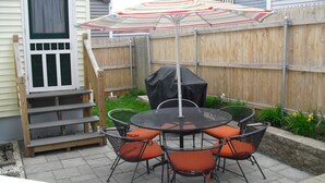 Une terrasse avec une table ronde et des chaises, un barbecue couvert et une clôture en bois.