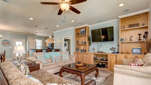 A spacious living room with a ceiling fan, a large flat-screen TV, and a wooden cabinet with decorative items. - Beautiful Gulf Front home at the waves - Southern Breeze (Dauphin Island)