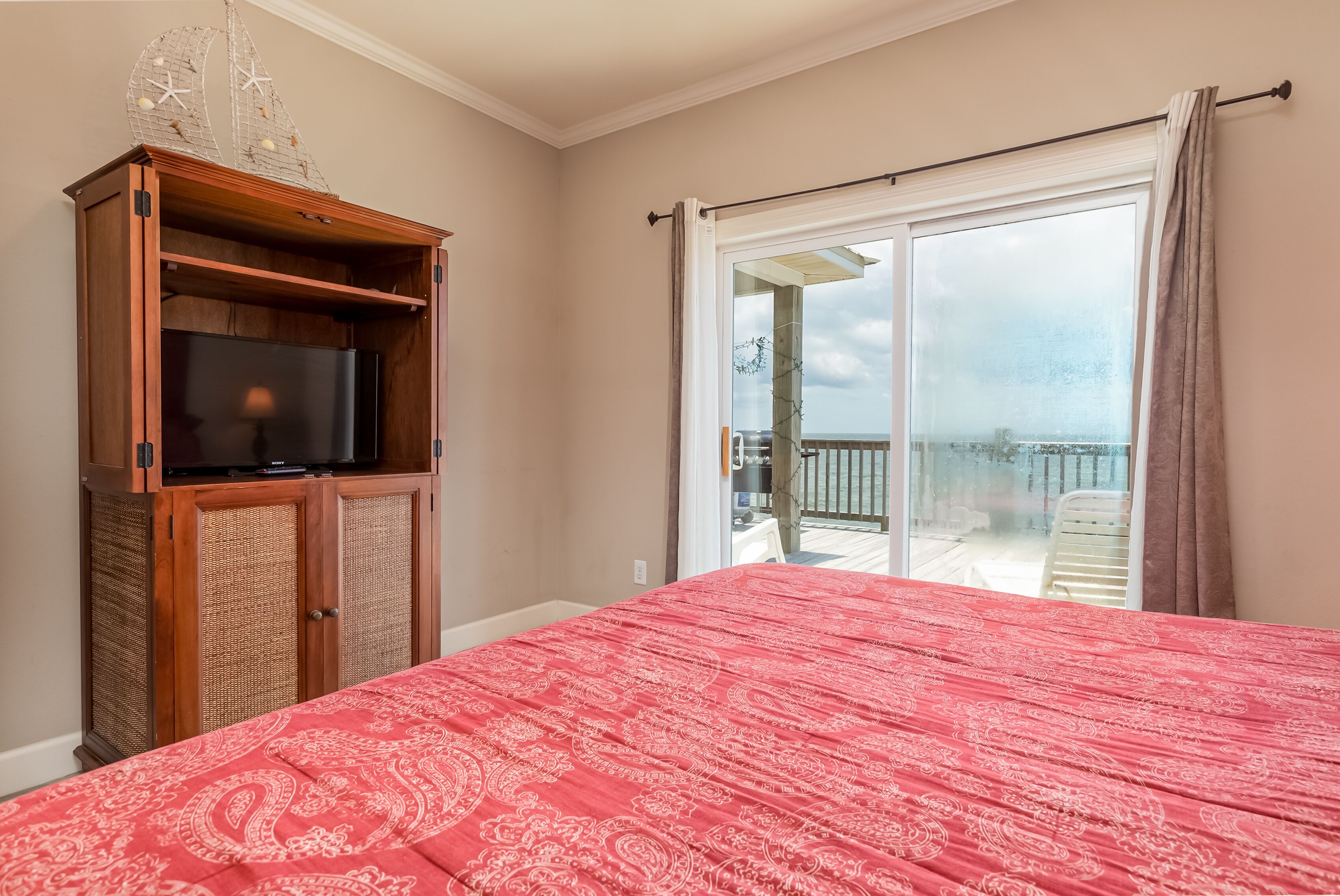 A bedroom with a red patterned bedspread, a wooden TV cabinet, and a sliding glass door leading to a balcony.