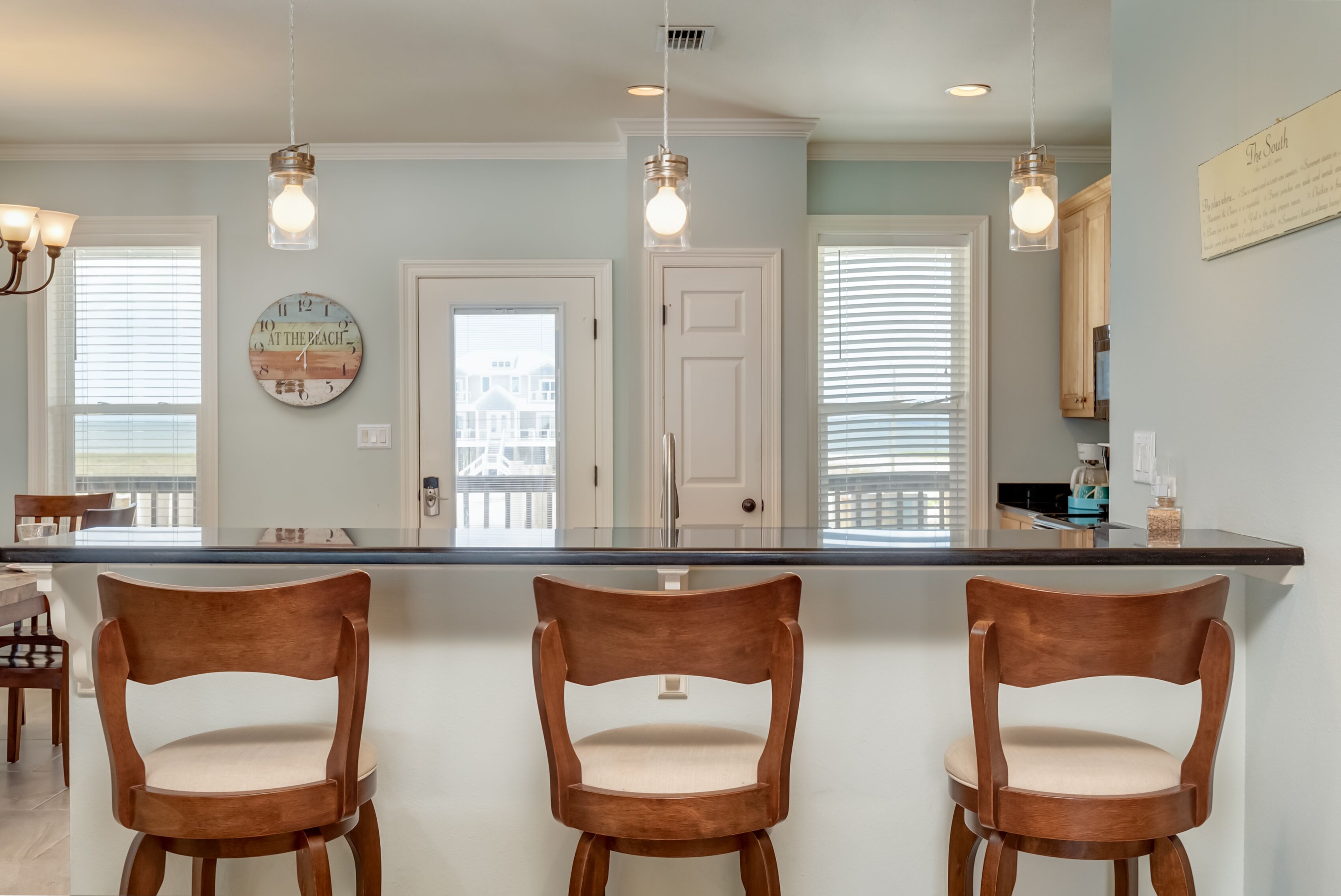 A kitchen with a central island, wooden chairs, pendant lights, and a view of the outside.