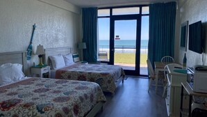 Hotelkamer met twee bedden, een bureau, een stoel, een televisie en een balkon met uitzicht op het strand.