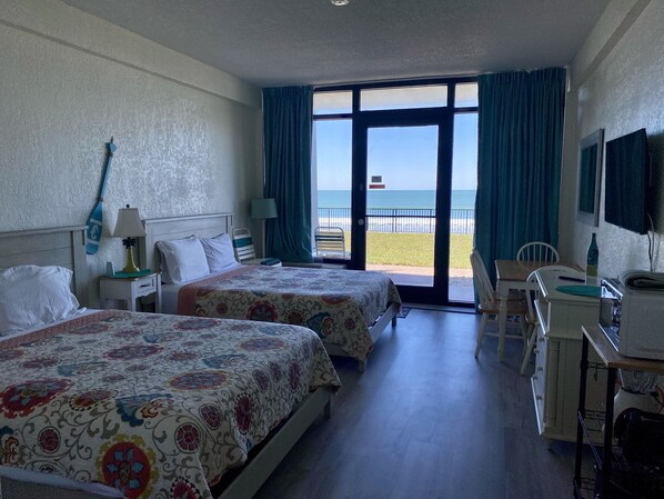 A hotel room with two beds, a desk, a chair, a TV, and a balcony with a view of the beach. - Oceanfront Studio 1st floor- Pools Open - Hawaiian Inn (Daytona Beach Shores)