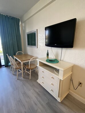 A room with a wooden dining table, white chairs, a white cabinet with drawers, a mounted flat-screen TV, and a window with green curtains. - Oceanfront Studio 1st floor- Pools Open - Hawaiian Inn (Daytona Beach Shores)