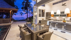 Een terras met een glazen tafel en rieten stoelen, een keuken met moderne apparatuur, en een uitzicht op de avondlucht.