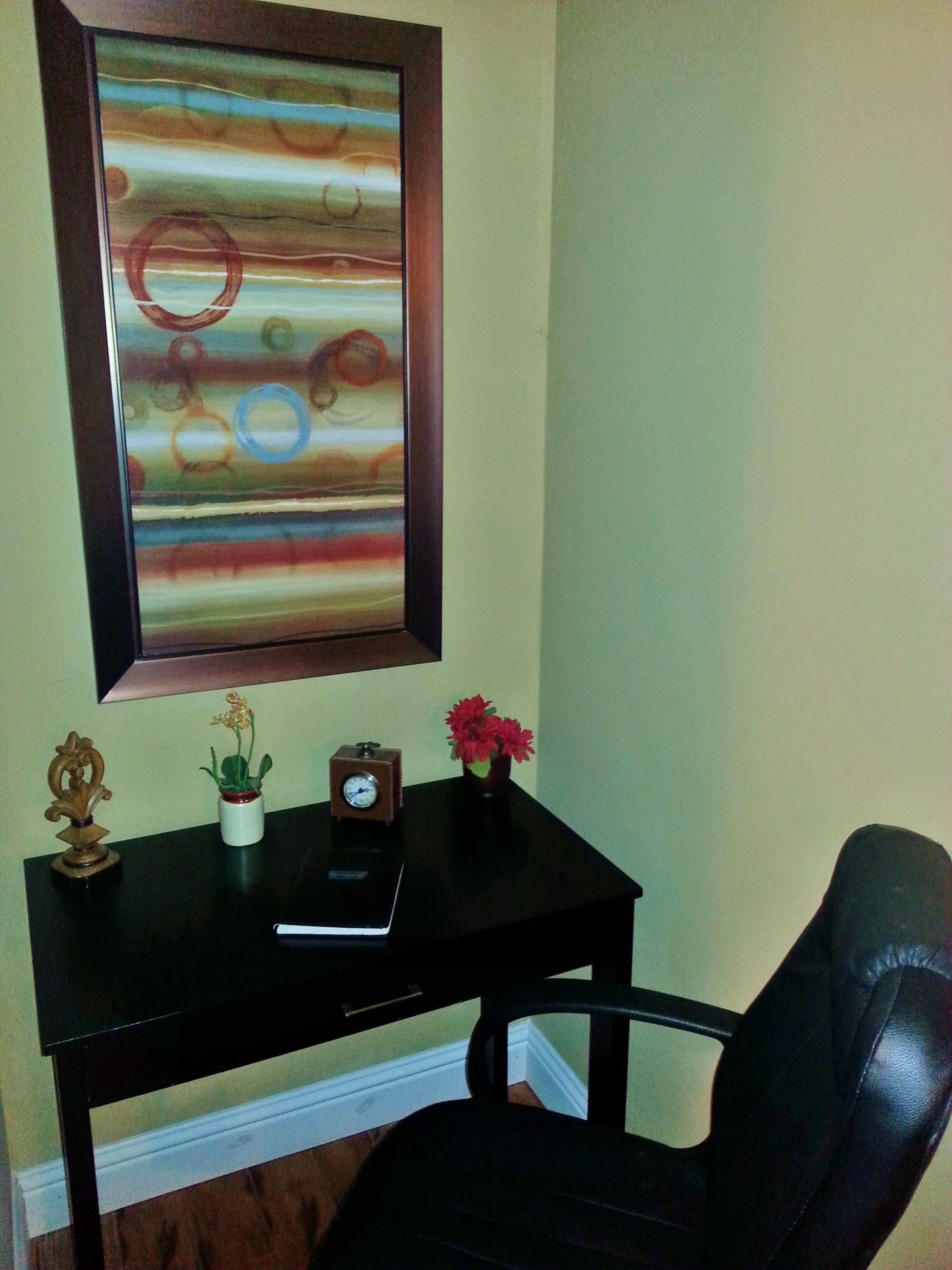 A room with a black desk, a chair, a clock, a vase with flowers, and a framed abstract painting on the wall.