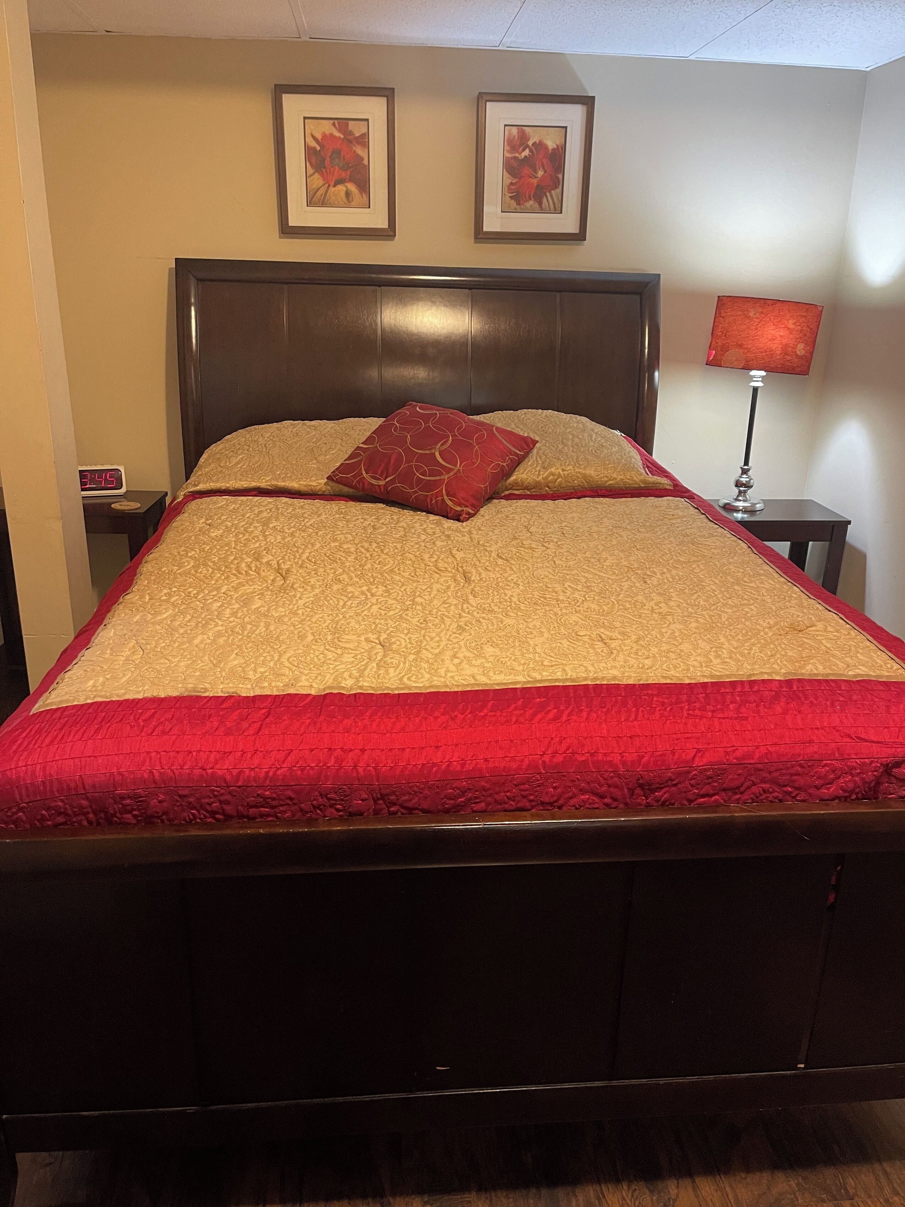 Un lit avec une couvre-lit rouge et beige, une tête de lit en bois et deux tableaux encadrés accrochés au mur.