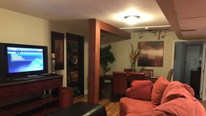 Una sala de estar con un sofá rojo, un reposapiés negro, un televisor, una mesa de comedor con sillas y un reloj de pared.