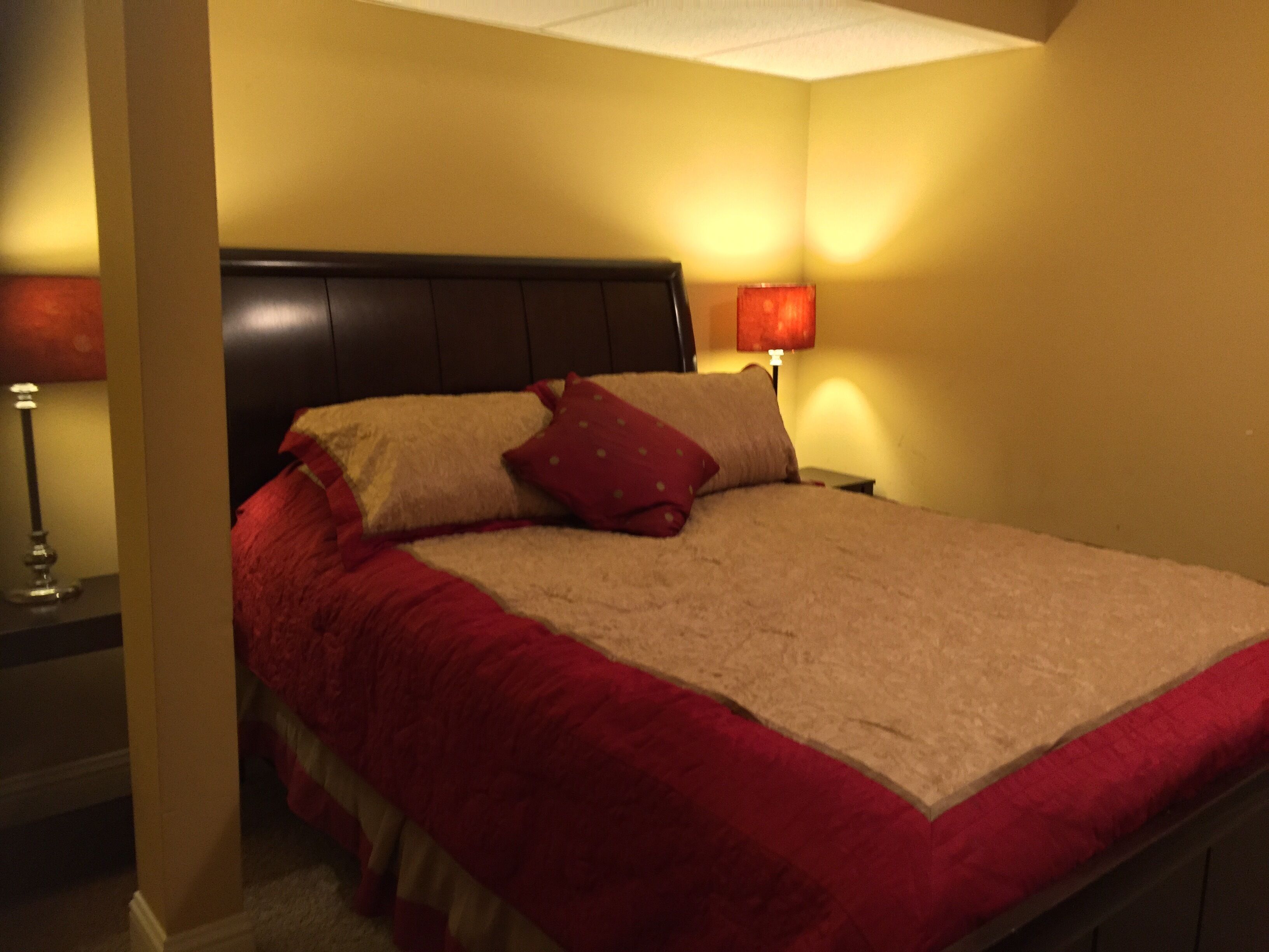 Una cama bien hecha con una colcha roja y beige, dos almohadas y un cabecero. Hay dos lámparas de noche a cada lado de la cama.