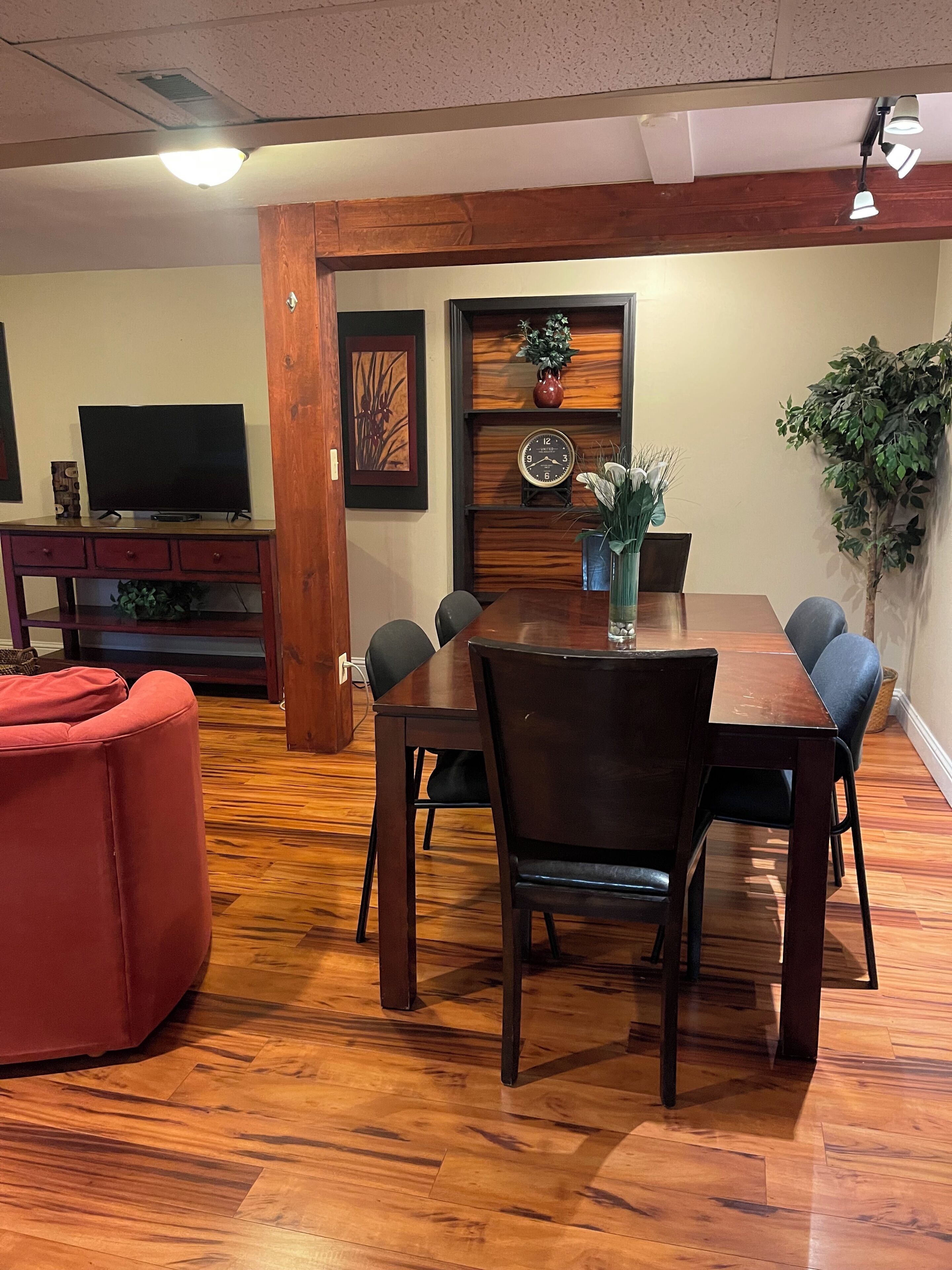 A dining area with a wooden table and chairs, a television on a stand, and a red sofa.
