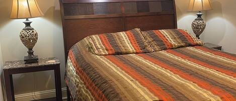 Un dormitorio con cabecera de madera, colcha a rayas, dos lámparas fijadas a la pared y una mesita de noche con una lámpara.
