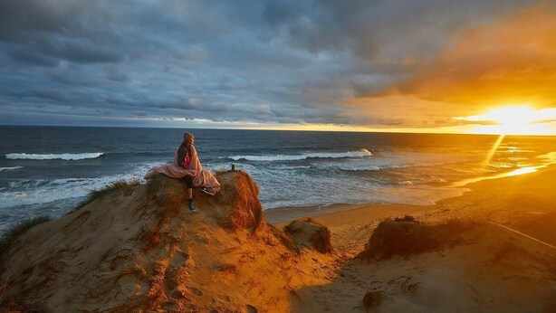 Une personne assise sur une plage de sable, avec vue sur l’océan et un coucher de soleil en arrière-plan.