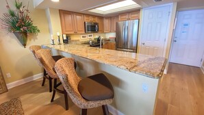 A modern kitchen with a central island, wooden bar stools, stainless steel appliances, and a hanging plant. - Sanibel Harbour Resort732: Experience dolphin tours by day and sunset views from your private lanai. (Fort Myers)