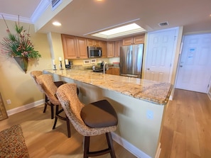 A modern kitchen with a central island, wooden bar stools, stainless steel appliances, and a hanging plant.