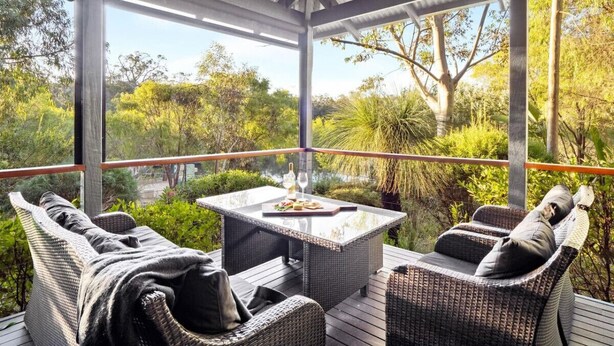 A covered outdoor seating area with wicker furniture and a table set for two, overlooking a lush garden. - Abundance | Sophisticated Luxury | River View (Margaret River)