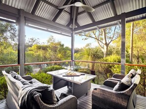 A covered outdoor seating area with wicker furniture and a table set for two, overlooking a lush garden.