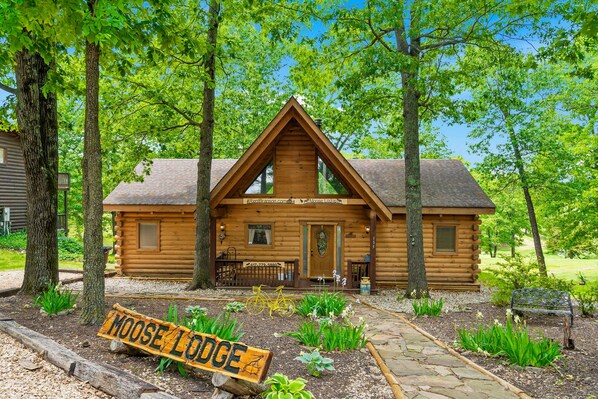 A log cabin with a triangular roof, a front porch, and a sign that reads 'MOOSE LODGE.'