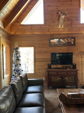Ein gemütliches Blockhaus-Interieur mit Holzbalkendecke, einem aufgehängten Hirschgeweih, einem Weihnachtsbaum, einem Ledersofa, einem Fernseher und einem Holzschrank.