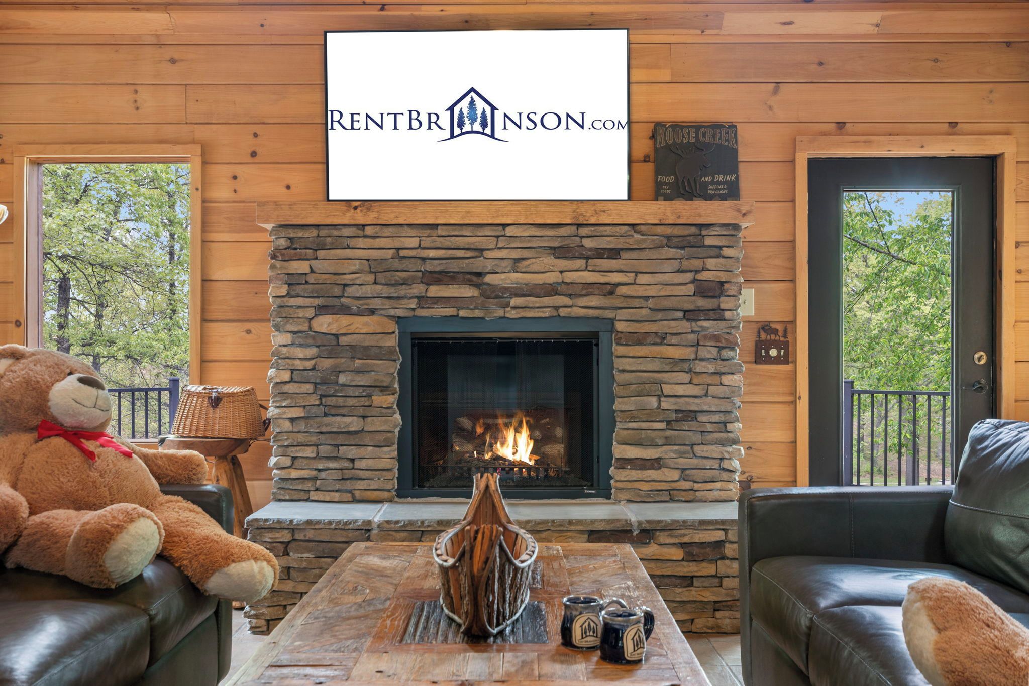 A cozy living room with a stone fireplace, a large teddy bear, and a sign for RENTBRINSON.COM.