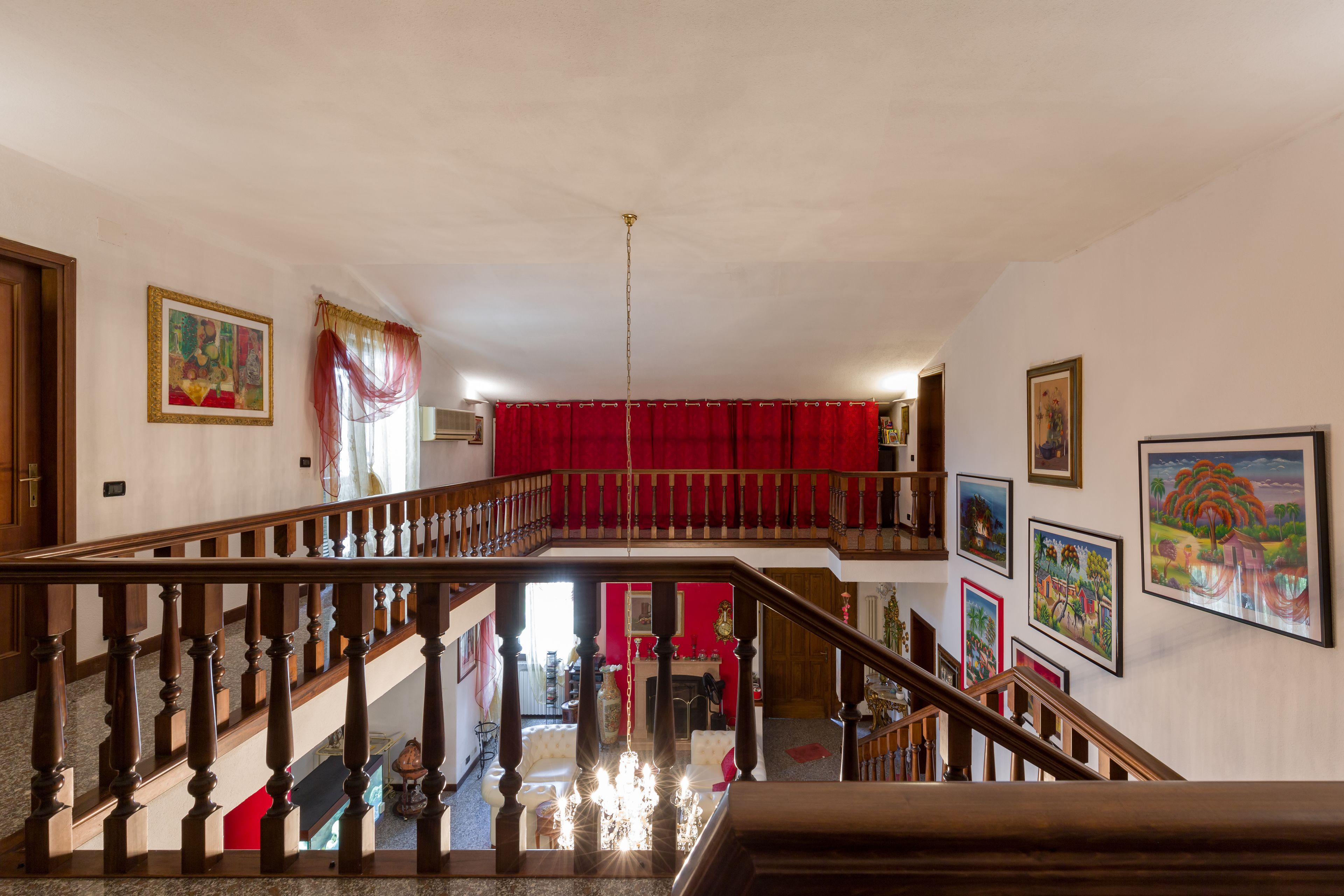 A multi-level interior with a wooden staircase, red curtains, and various framed artworks on the walls.