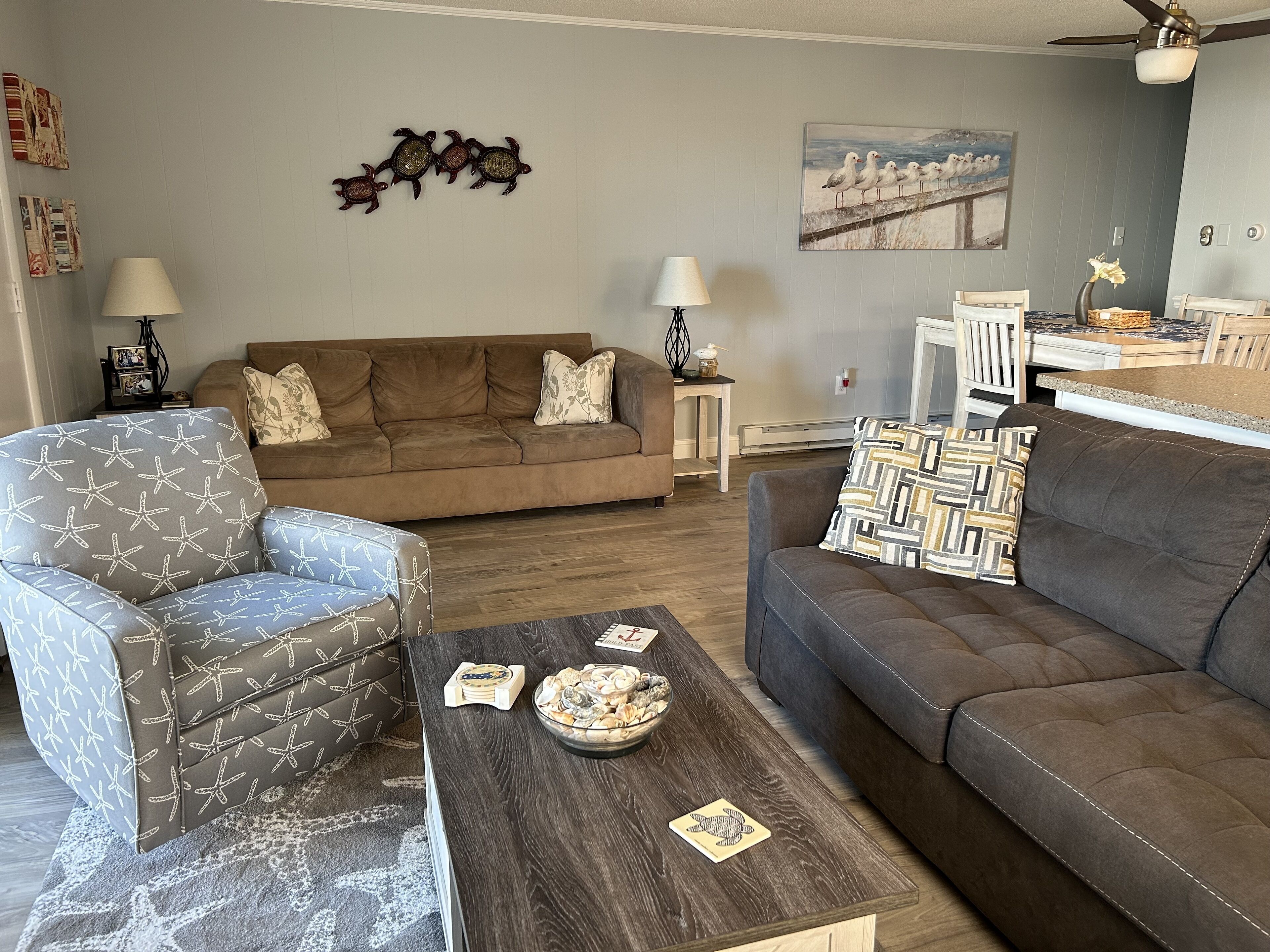 A living room with a brown sofa, a patterned armchair, a coffee table with decorative items, and wall art depicting seagulls and turtles.