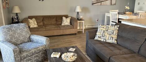 A living room with a brown sofa, a patterned armchair, a coffee table with decorative items, and wall art depicting seagulls and turtles.