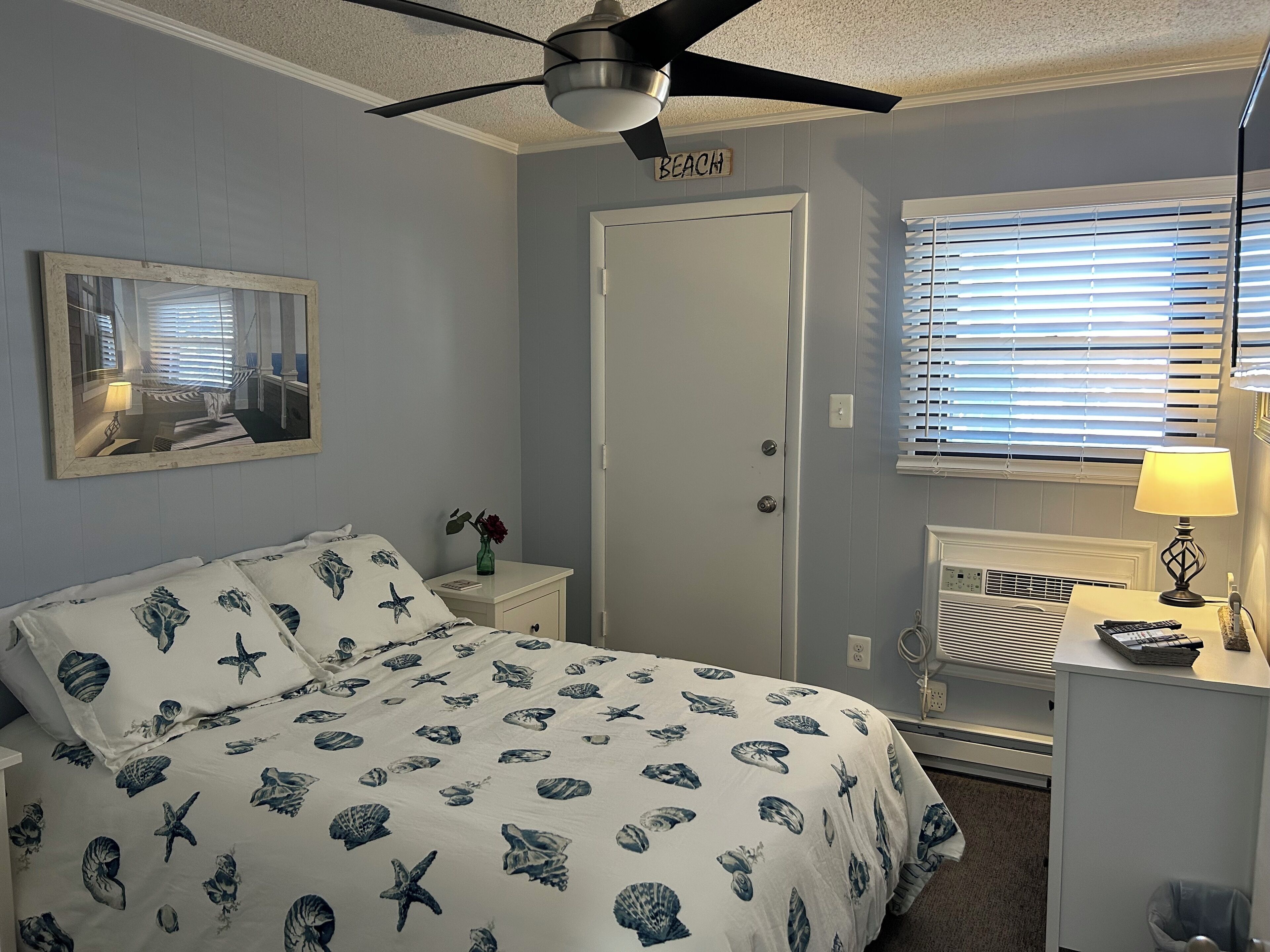 Ein Schlafzimmer mit einem Bett, einem Nachttisch, einem Spiegel, einer Tür mit der Aufschrift 'BEACH', einem Fenster mit Jalousien und einem Deckenventilator.