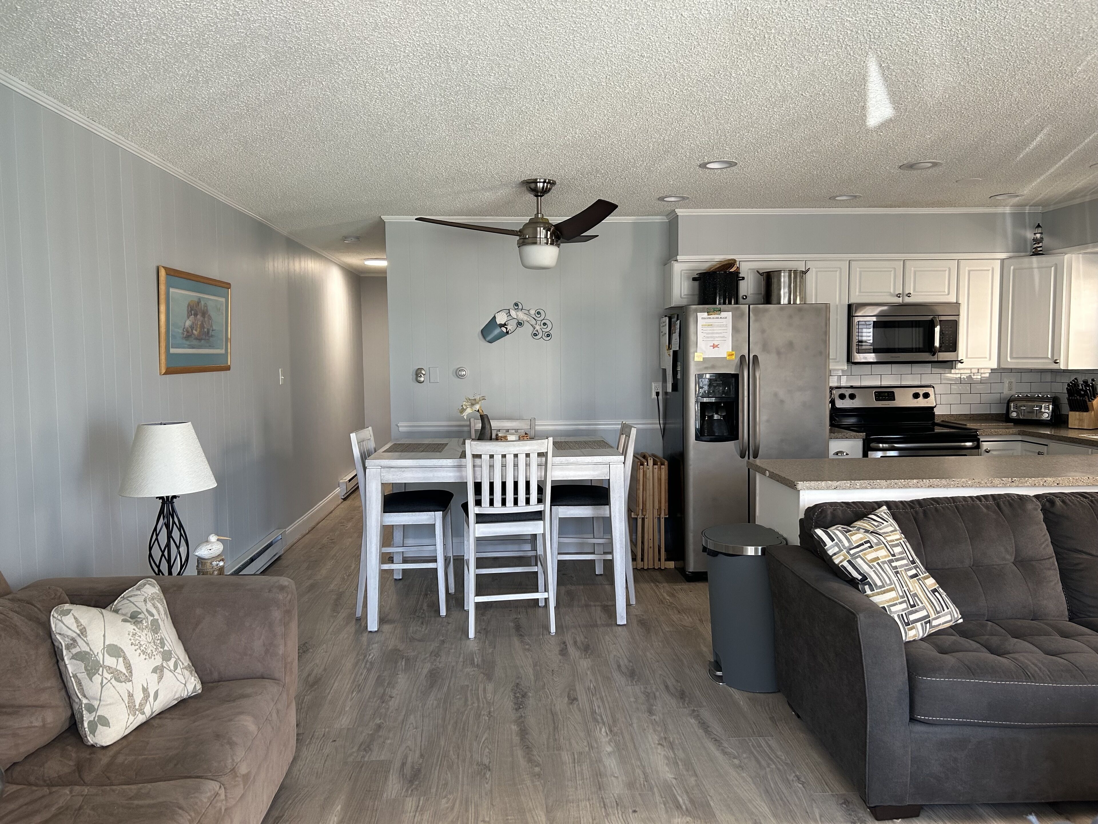 A modern kitchen with white cabinets, a stainless steel refrigerator, and a microwave. There is a dining table with chairs and a ceiling fan above.