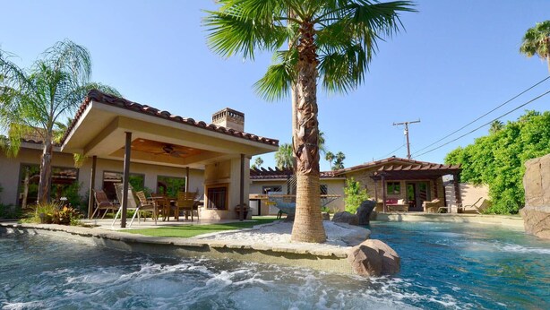 A pool with a waterfall feature, surrounded by a house with a covered patio and outdoor seating. - LAZY RIVER!  SLIDE and WATERFALLS!  GAMEROOM. (Palm Springs)