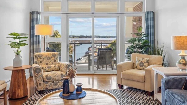 A spacious living room with a large sliding glass door leading to a balcony with a view of the water. There are two armchairs, a coffee table, and a lamp. - Luxury Waterfront Marina Condo in Downtown (Charlottetown)